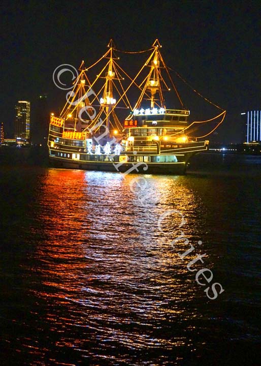 A brightly lit, masted brig sails up and down the Huangpu River in front of the Bund and Pudong.